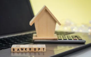 Toy house on calculator atop laptop, with wooden blocks spelling BANKRUPT, illustrating financial distress and risk of eviction