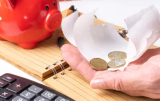 Red piggy bank with hand holding rolled coins, notebook, pen, and calculator on desk, symbolizing saving, budgeting, and asset protection