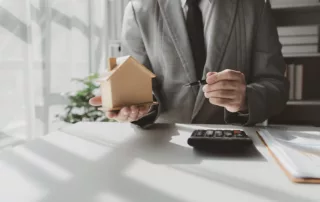 Man in a suit holding a small wooden house model and a pen over documents and a calculator on a desk — representing real estate, homeownership, and decisions about keeping a home during Chapter 7 bankruptcy.