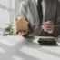 Man in a suit holding a small wooden house model and a pen over documents and a calculator on a desk — representing real estate, homeownership, and decisions about keeping a home during Chapter 7 bankruptcy.