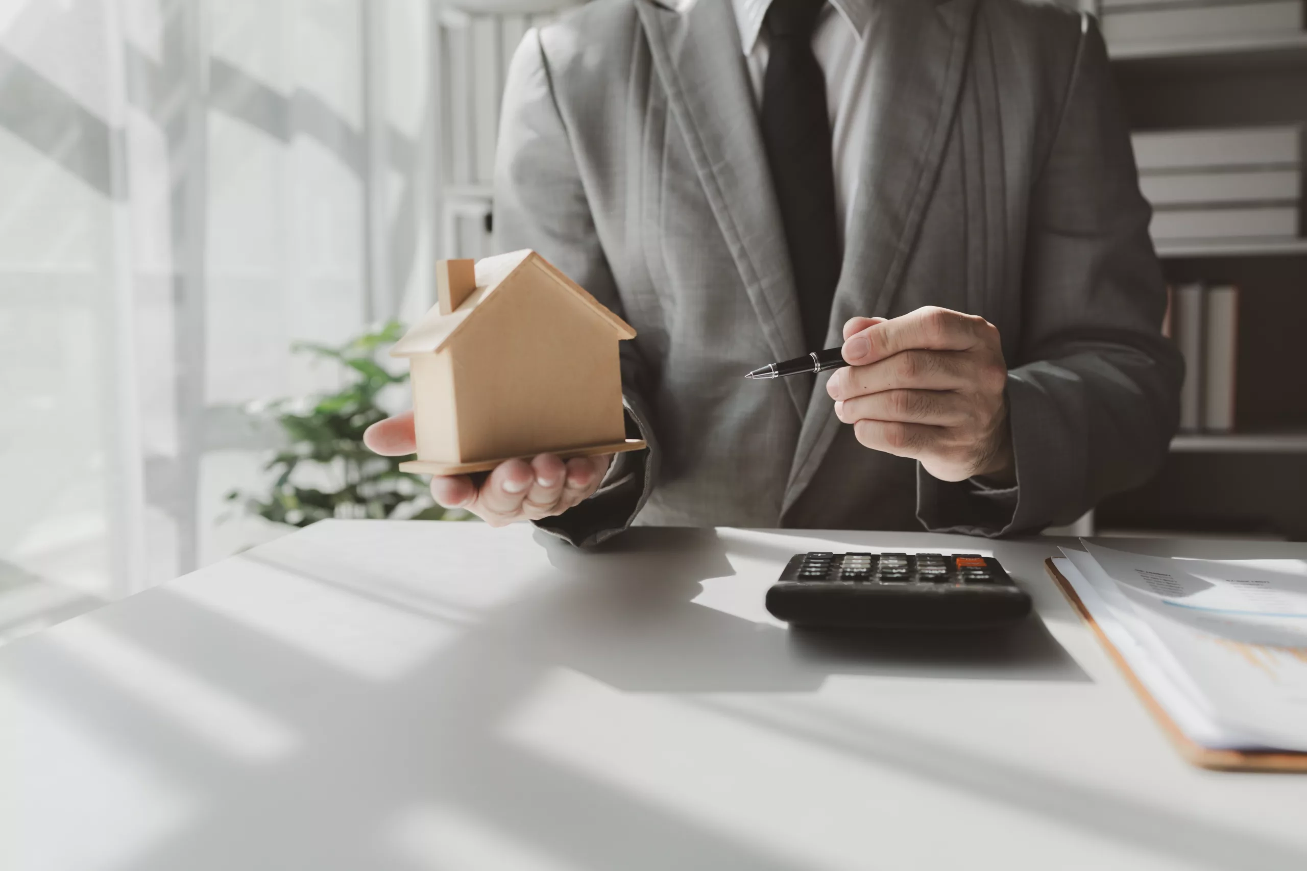 Man in a suit holding a small wooden house model and a pen over documents and a calculator on a desk — representing real estate, homeownership, and decisions about keeping a home during Chapter 7 bankruptcy.